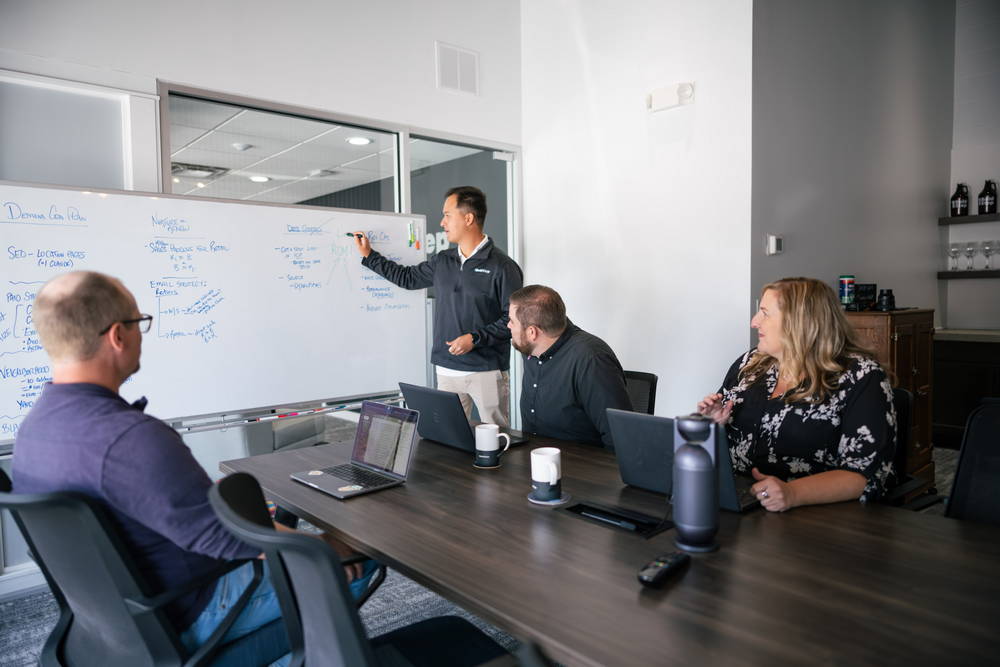 Four marketing professionals in a modern conference room collaborating around laptops and a whiteboard.