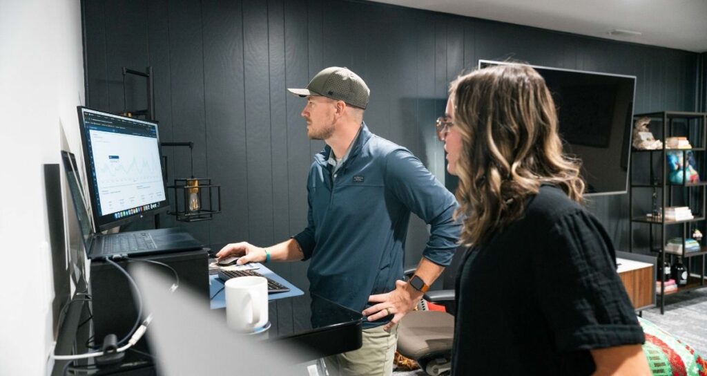 Two 1BoldStep team members collaborating at a laptop in an office with black wood panel walls