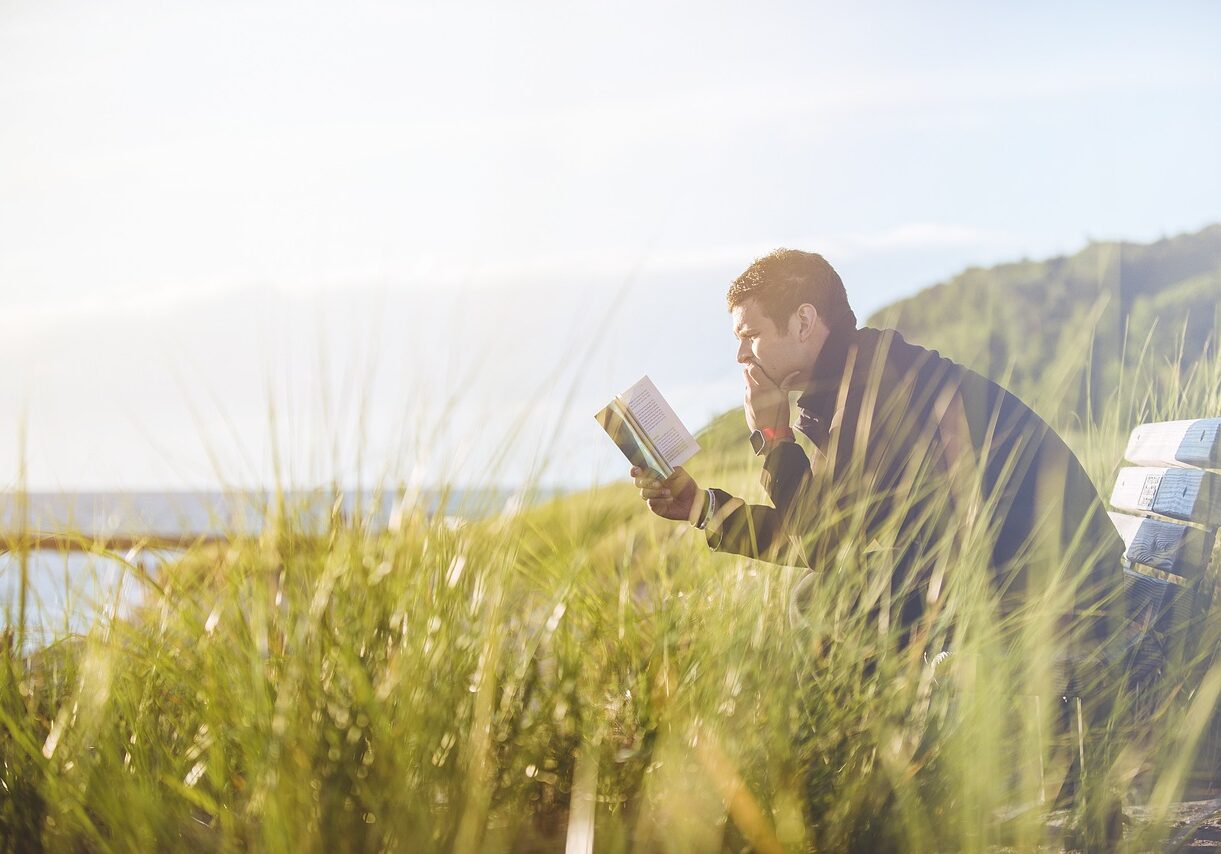 man reading on a bench on a hill next to a body of water
